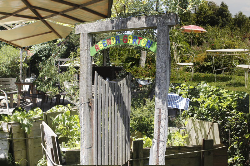 Das Eingangstor mit dem Schriftzug "Agendagarten" in bunten Buchstaben, dahinter sieht man ein Hochbeet mit Salat, Markisen und einen Holztisch, sowie auf einer Wiese Gartenstühle und Sonnenschirme.