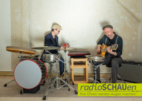 Zwei Musiker sitzen vor einer Wand, links am Schlagzeug, rechts mit einer Gitarre